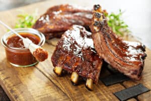 rack of ribs on a wooden cutting board sitting next to a cup of barbeque sauce and a baster